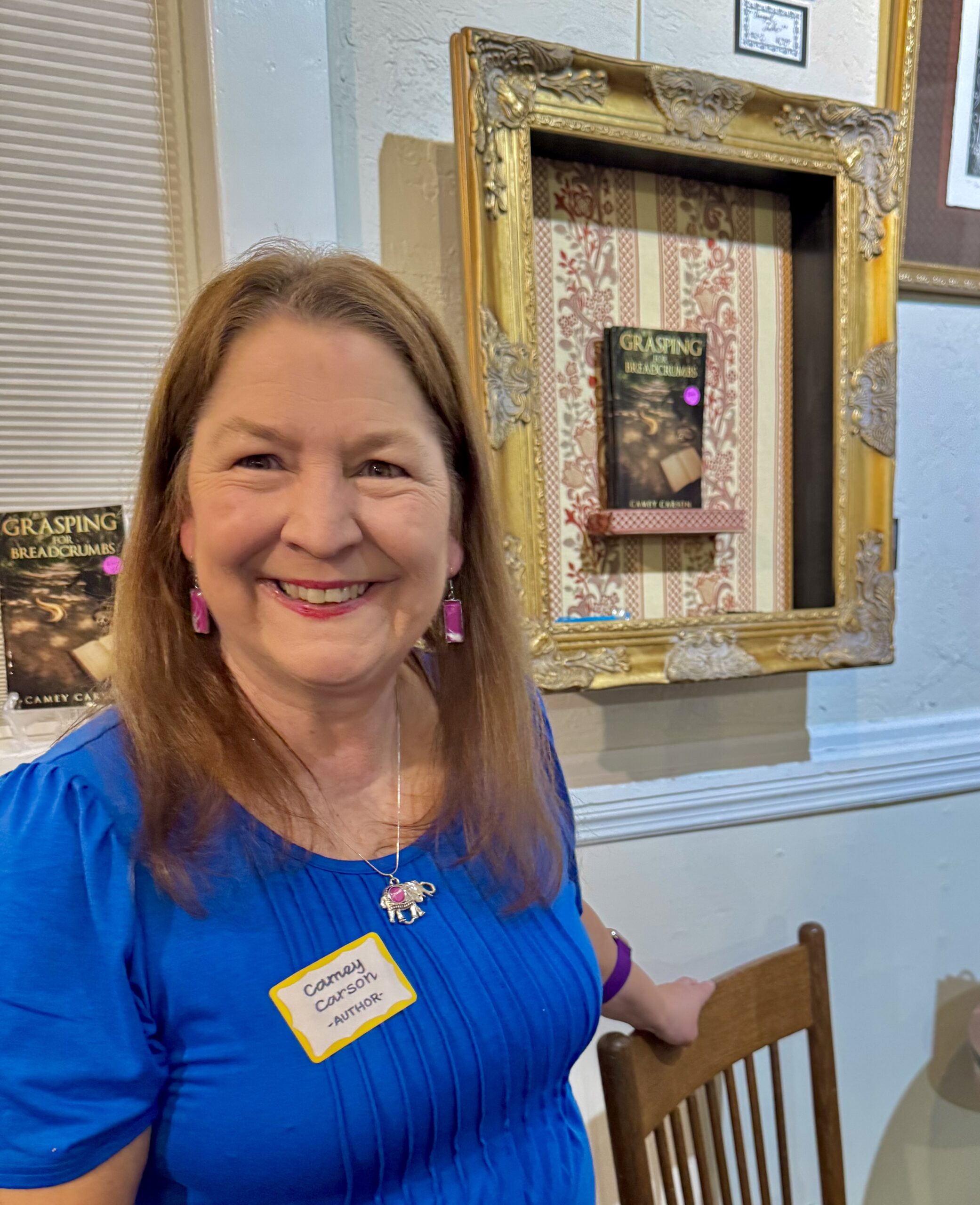 Picture of author Camey Carson at the Melrose Artwalk 11/7/25 standing in front of a framed copy of her first, fiction release "Grasping for Breadcrumbs"
